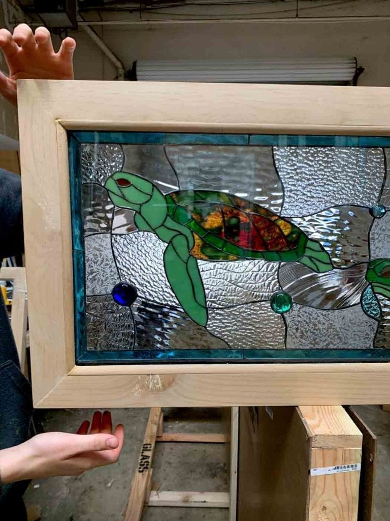 A rectangular stained glass panel in a light natural wood frame, featuring a vibrant green sea turtle with a multi-colored shell (red, orange, yellow) swimming in clear, textured glass water. The panel is accented with round blue and green glass jewels and bordered by teal glass, with a second turtle partially visible on the right.