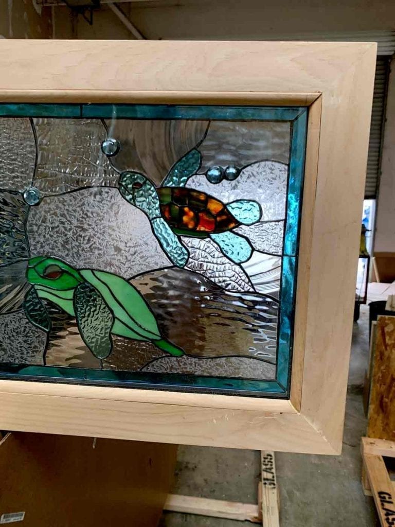 A leaded stained glass panel showcasing two sea turtles swimming. One turtle is vibrant green, and the other features a multi-colored orange and brown shell with blue-green flippers. They are surrounded by various clear textured glass types resembling water and several blue glass bubbles. The panel is framed in light natural wood.