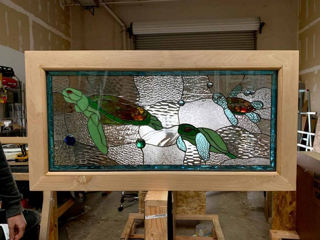A rectangular stained glass panel featuring three green sea turtles swimming amidst clear, textured glass representing water and bubbles, all set within a light natural wood frame.
