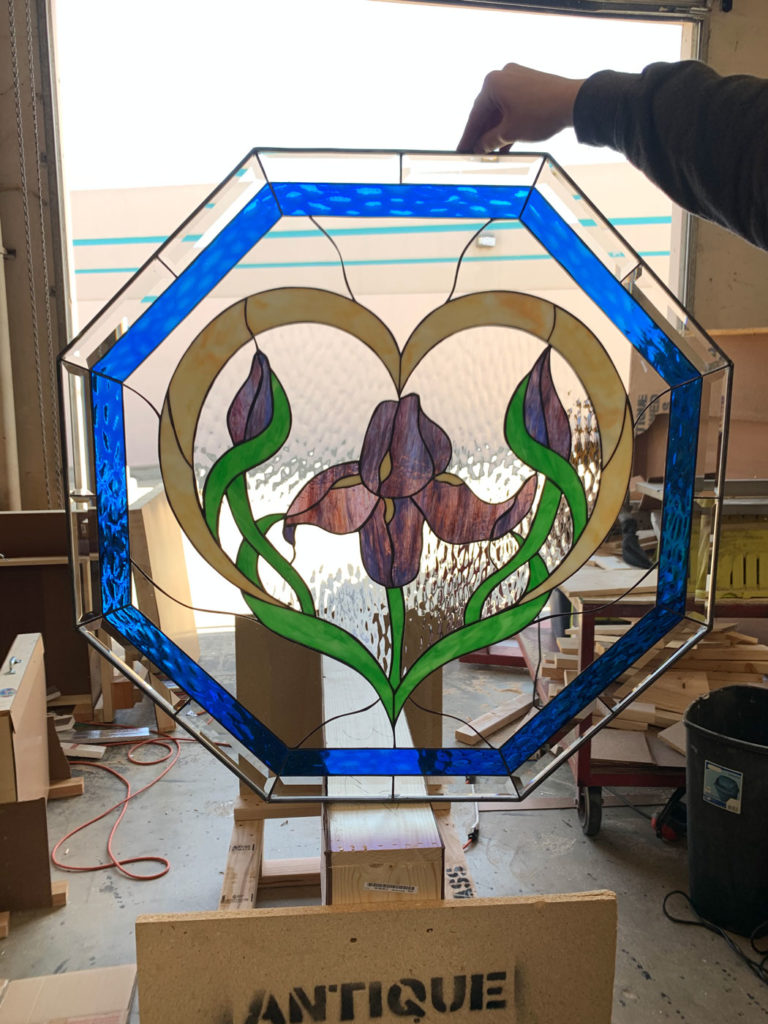 An octagonal leaded stained glass panel featuring a central iris flower in purple and brown hues with green stems and buds, framed by a golden heart shape against a clear textured background, all bordered by vibrant blue glass.