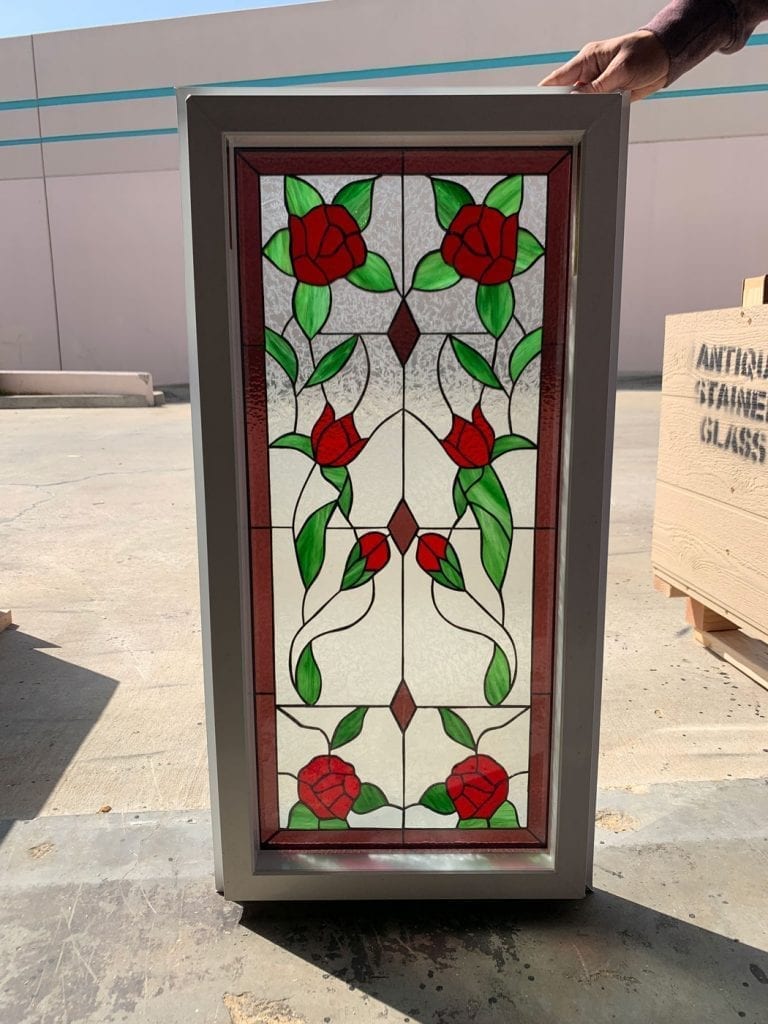 A rectangular stained glass window panel in a modern gray frame, showcasing a symmetrical floral design with vibrant red roses, green leaves, and winding stems, set against a clear textured background, and bordered by dark reddish-brown glass.