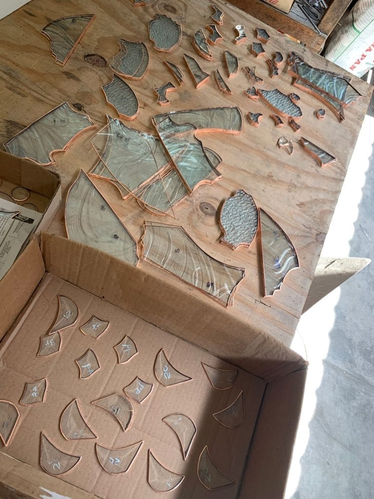 Numerous cut glass pieces for a stained glass project, featuring clear textured and swirled glass, meticulously wrapped in copper foil and laid out on a wooden workbench. Smaller, numbered foiled pieces are organized in a cardboard box.