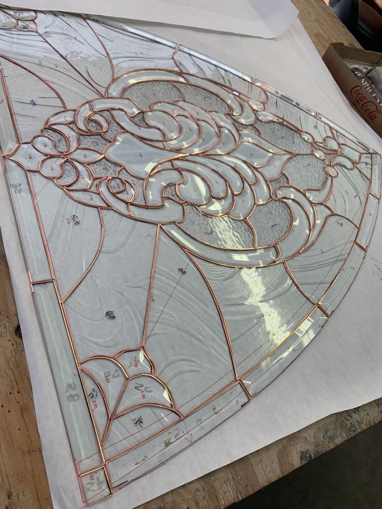 A semicircular stained glass panel in the midst of assembly using the copper foil (Tiffany) method. The intricate design features organic, swirling patterns composed of various clear textured glass pieces, including hammered and streaky textures, all meticulously wrapped in copper foil and laid out on a paper pattern on a wooden workbench.