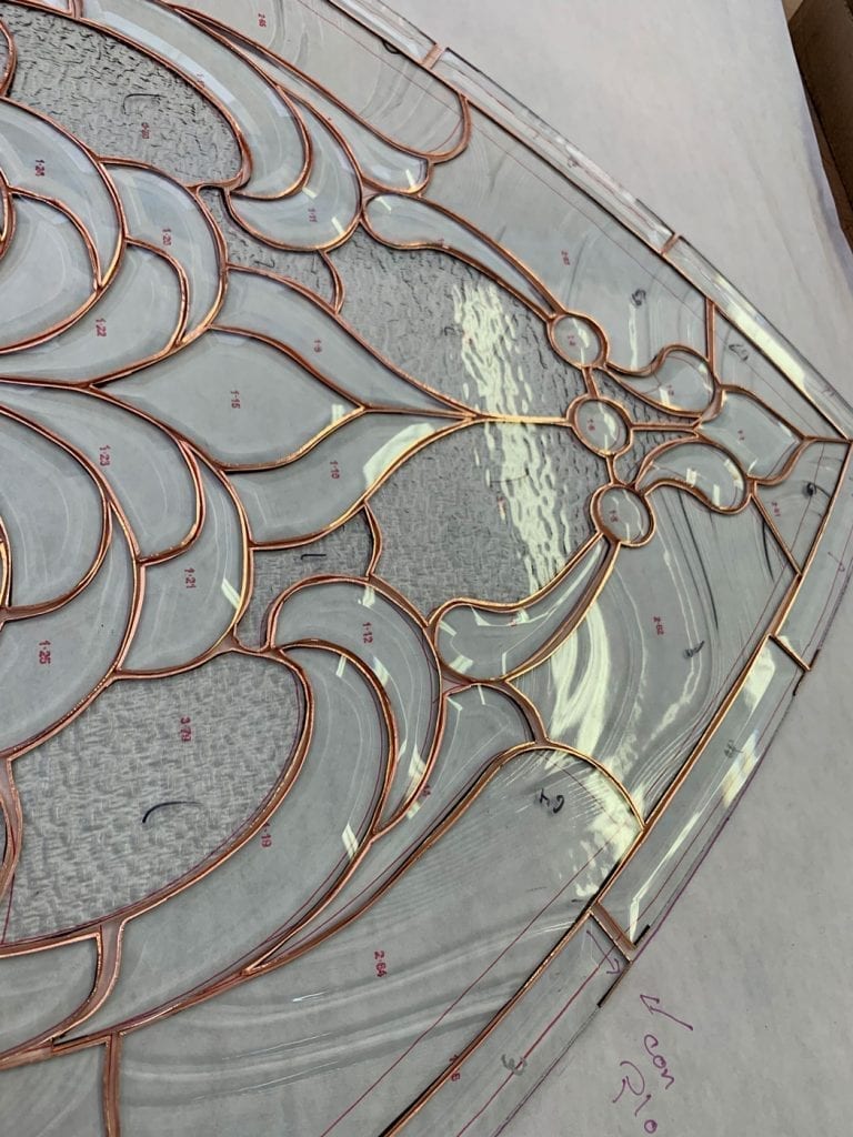 A close-up, overhead view of a stained glass panel in the making, featuring a flowing, organic Art Nouveau style design. Individual pieces of clear textured and smooth glass are laid out on a paper pattern, each edged with shiny copper foil, ready for soldering. The design includes swirling shapes and a central section of rippled or water glass, with various glass pieces numbered for assembly.