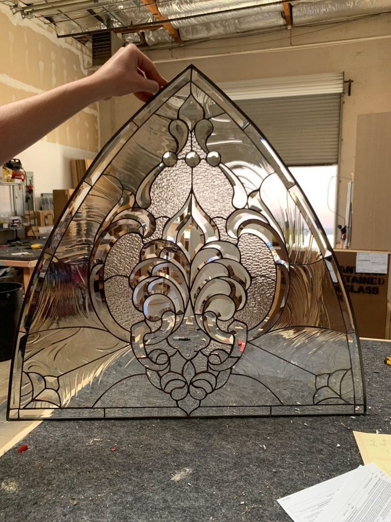 An elegant arched, leaded stained glass transom panel featuring a large central Fleur-de-lis motif, surrounded by clear, textured, and geometrically arranged beveled glass, reflecting light in a workshop setting.