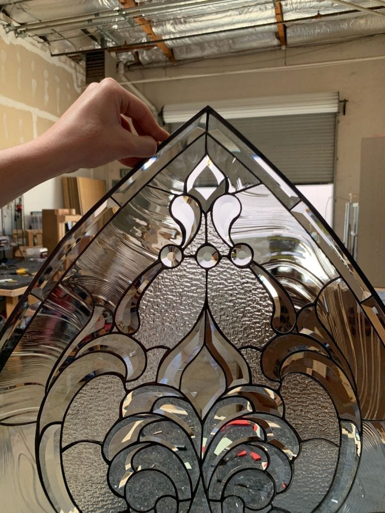Close-up of a hand holding an intricate clear beveled stained glass transom panel featuring scrollwork, fleur-de-lis elements, and various glass textures, with a workshop in the blurred background.