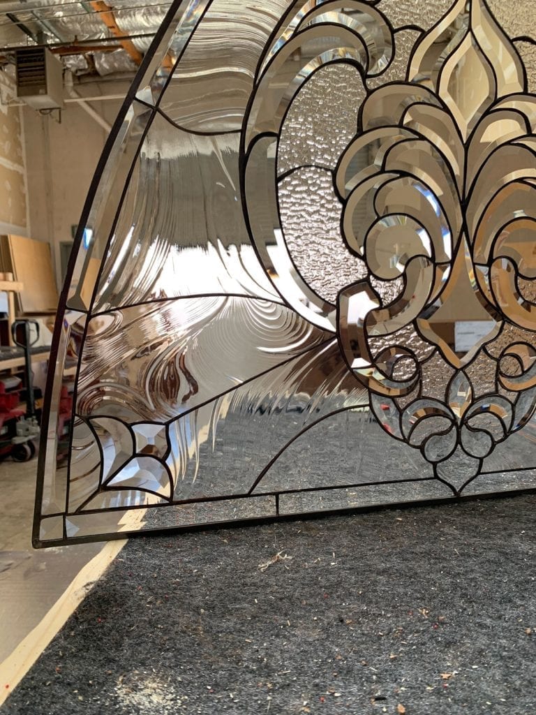 An ornate clear leaded stained glass panel showcasing a large central beveled Fleur-de-lis motif, surrounded by intricate scrolling patterns, and crafted from various clear textured and smooth glass pieces.