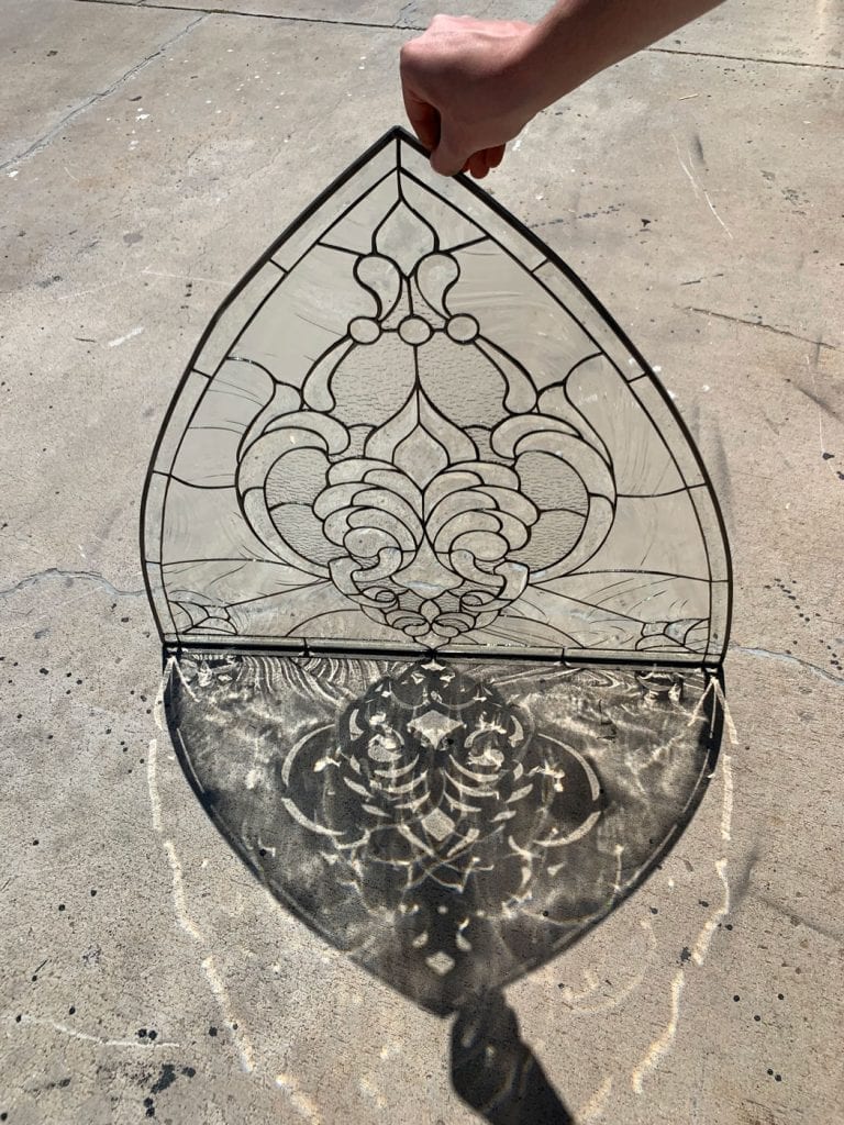 A human hand holds up an ornate clear leaded stained glass panel with a gothic arch shape, casting a dramatic, intricate shadow of its scrollwork and patterned glass onto a textured concrete ground under bright sunlight.
