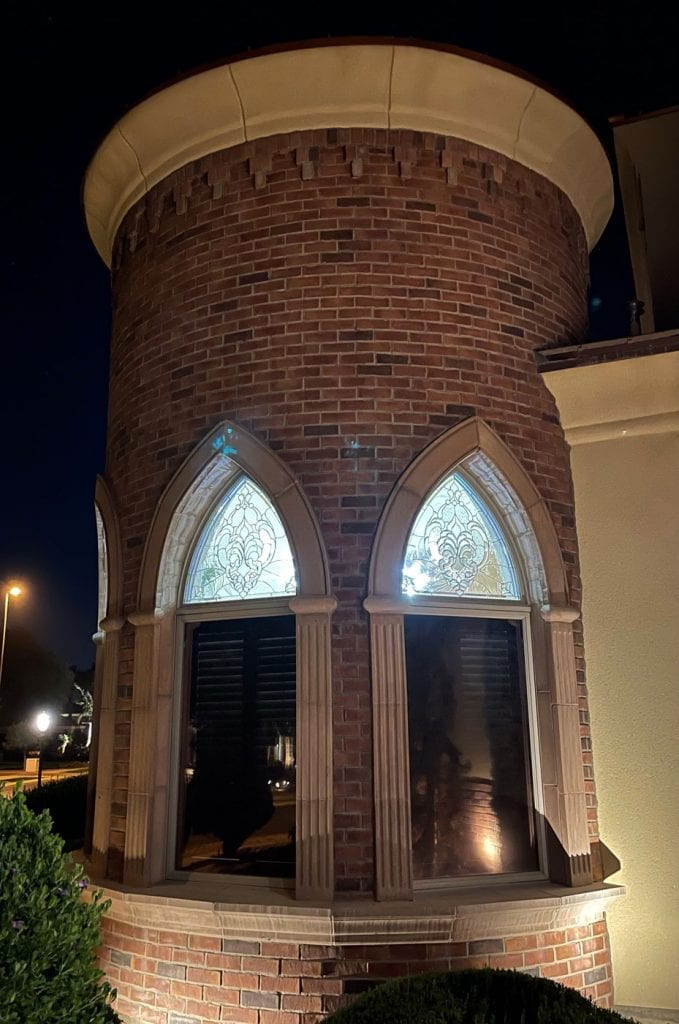 Two custom leaded stained glass arch transoms with an ornate clear scrollwork and Fleur-de-lis pattern, installed in a brick turret at night. The stained glass sits above dark lower window panes, framed by light-colored stone columns and arches, illuminated by ambient outdoor lighting.