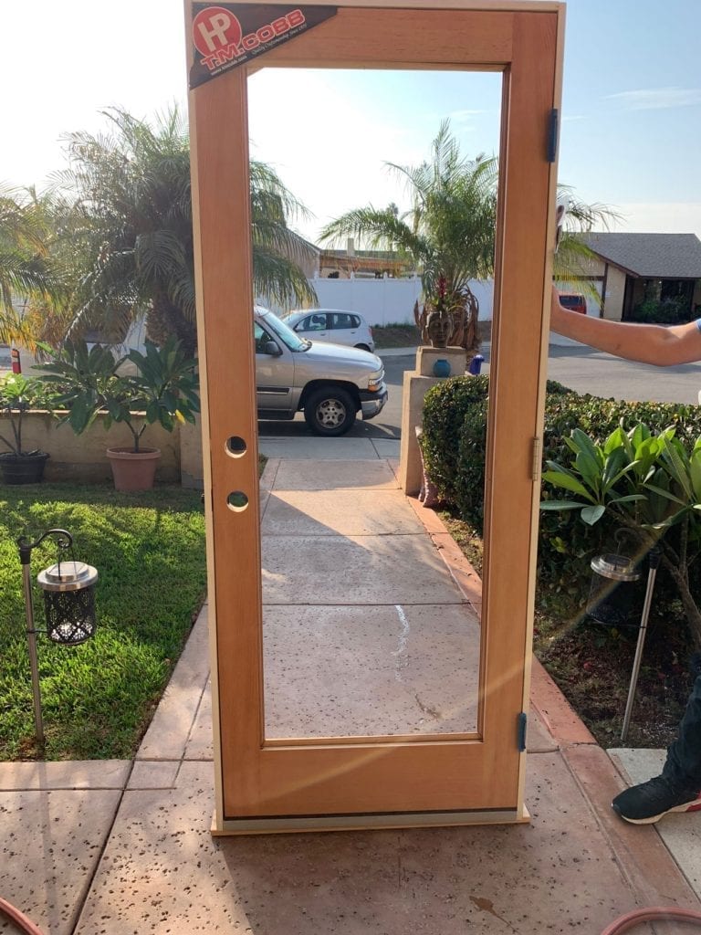 A new, unfinished T.M. Cobb full-lite wood entry door with clear glass, standing on a concrete walkway. The door features a natural light brown wood grain, black hinges, and pre-drilled holes for a doorknob and lock. A 'T.M. Cobb' brand sticker is visible in the top left corner of the door frame.