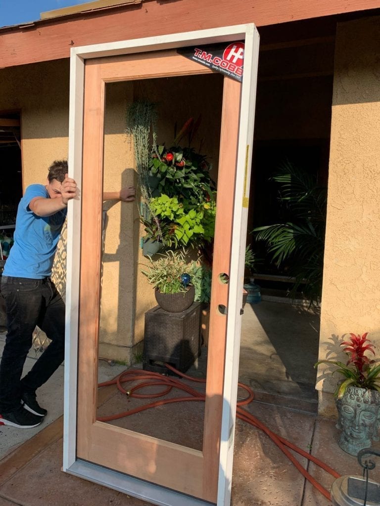 A man installs a pre-hung solid wood door frame with a large, clear glass panel. The light-colored wood door is framed by white casing, shown outdoors with plants and a beige stucco wall in the background.