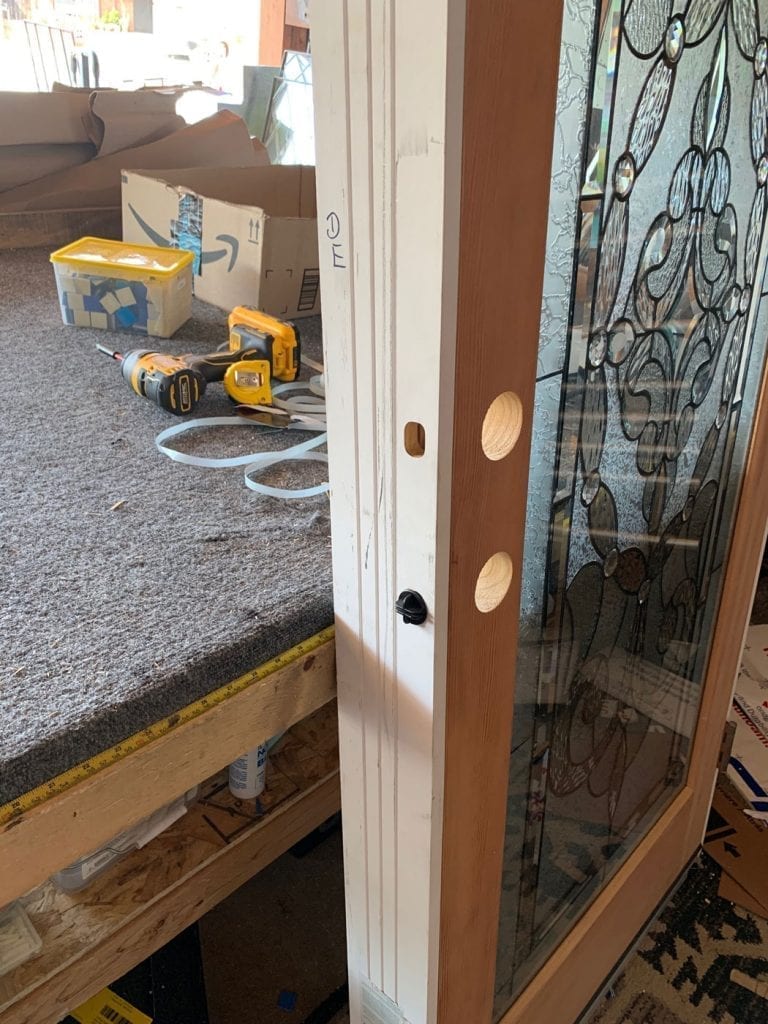 A pre-hung wooden entry door featuring an elegant clear beveled stained glass panel, shown mid-preparation for installation. The door jamb has precision-drilled holes for handle and lock hardware. On a workbench nearby, a drill, tape measure, and a container of small glass pieces are visible. The stained glass itself showcases a beautiful, organic pattern composed of clear, textured, and beveled glass elements.