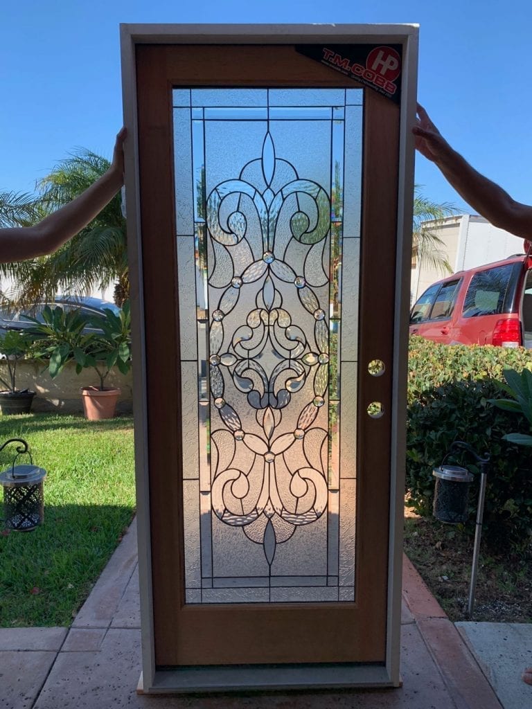 A full-lite exterior door with a light wood-toned frame, featuring a clear leaded stained glass panel. The panel showcases an intricate, symmetrical scrollwork and floral-inspired design, crafted with various clear textured and beveled glass pieces, accented by small circular clear jewels.