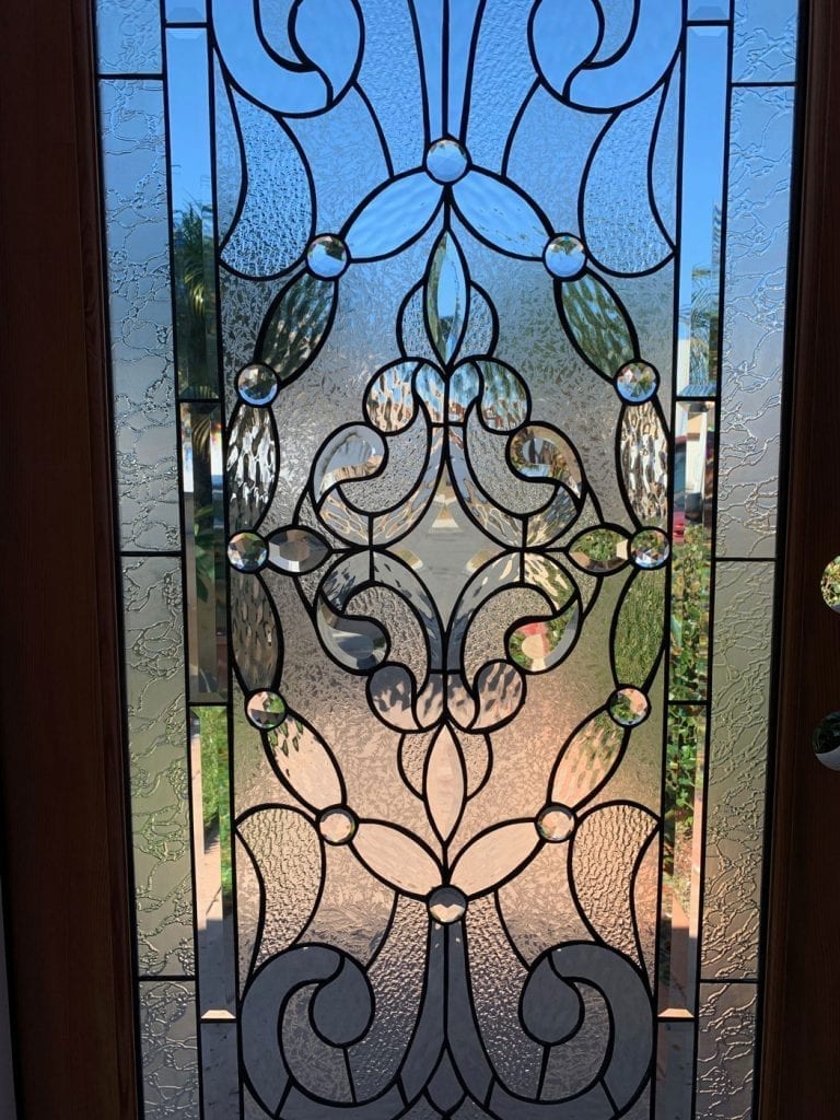 A clear leaded stained glass door panel featuring an intricate, symmetrical design of ornate scrolling motifs, stylized floral elements, and numerous round and oval clear bevels, all set within various textures of clear glass, framed by a dark caming.