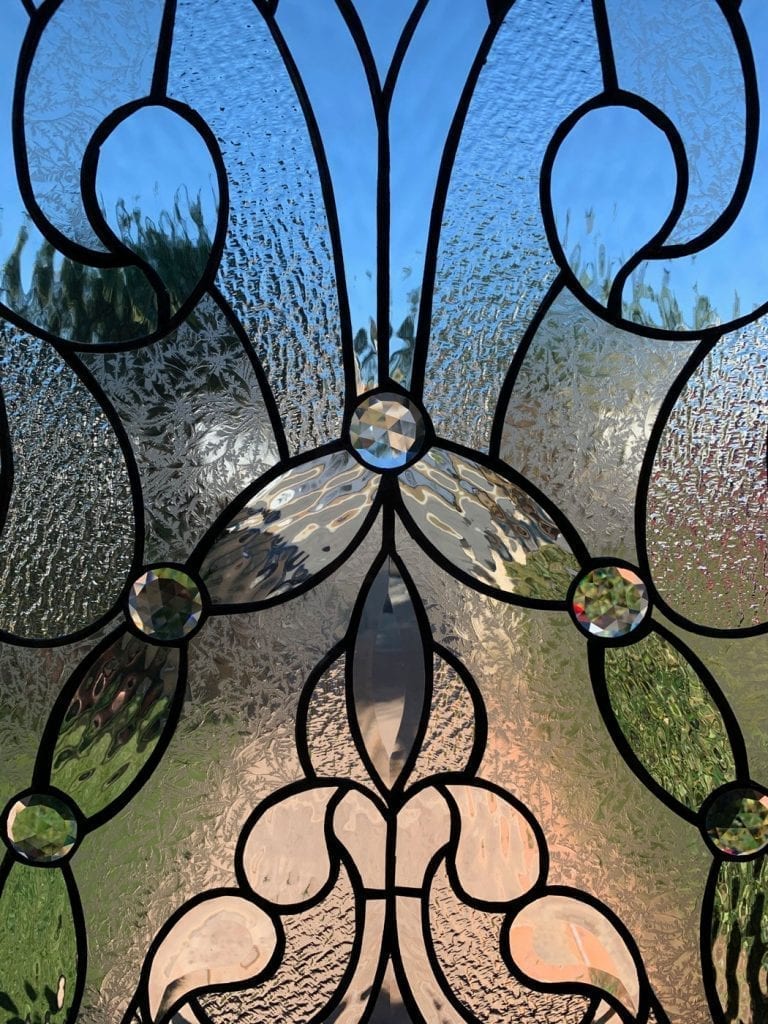 Close-up of a clear leaded stained glass panel featuring an intricate organic pattern of textured glass, clear beveled glass, and multiple faceted round jewels. Blurred blue sky and green foliage are visible through the glass.