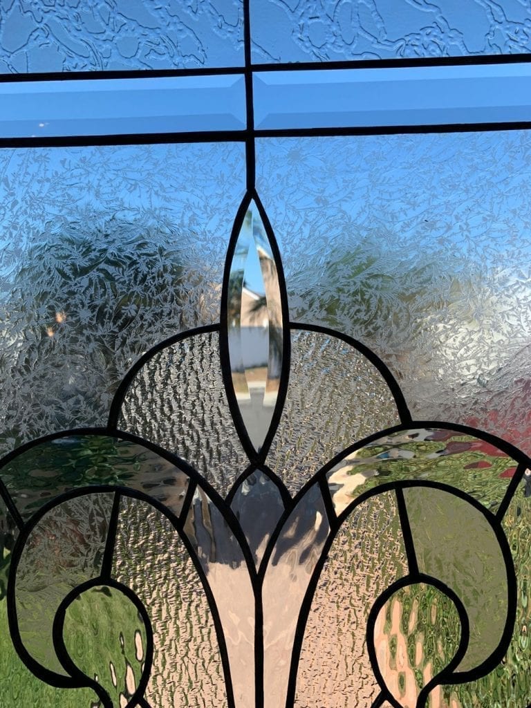 A close-up of a decorative leaded glass panel, showcasing an intricate design primarily with clear, textured, and beveled glass. A prominent, vertically oriented, teardrop-shaped beveled glass piece forms the center, surrounded by elegant scrolling patterns crafted from clear glue-chip textured glass. Rectangular clear beveled glass sections are visible at the top, reflecting the blue sky.