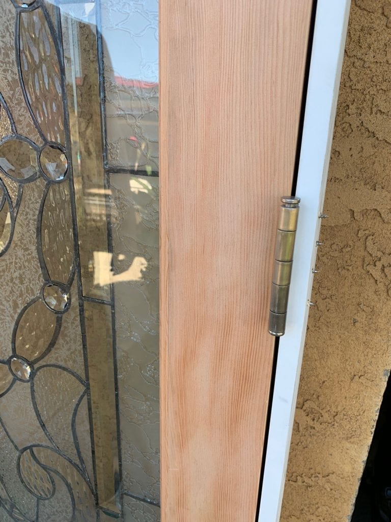 A close-up view of a door showcasing a clear, textured, and beveled stained glass insert on the left. The glass features a classic scroll and floral pattern outlined with dark lead lines, incorporating circular and oval bevels alongside clear textured background glass. To the right, a natural, light-toned wood panel with prominent vertical grain provides a warm contrast. A brass-colored door hinge is visible on the right edge, attaching the wooden section to a white door frame.
