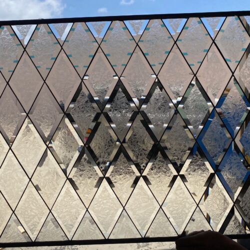 A rectangular leaded stained glass panel featuring a repeating diamond pattern. Alternate diamonds are made of clear beveled glass, while the others are clear textured glass with a frost-like or crackled appearance. Dark lead lines separate all the pieces, and a bright blue sky is visible through the panel.