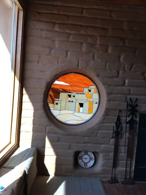 A circular stained glass window embedded in an adobe-style brick wall, depicting a stylized Southwestern pueblo village with light-colored buildings and a vivid orange and yellow sky. Sunlight streams from a large window on the left, casting sharp shadows across a light-colored couch and the textured wall.