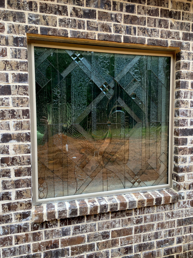 A rectangular clear leaded stained glass window installed in a dark brown brick wall. The window features an abstract geometric pattern with a variety of clear textured and beveled glass pieces. Trees and brown ground are visible distorted through the glass.