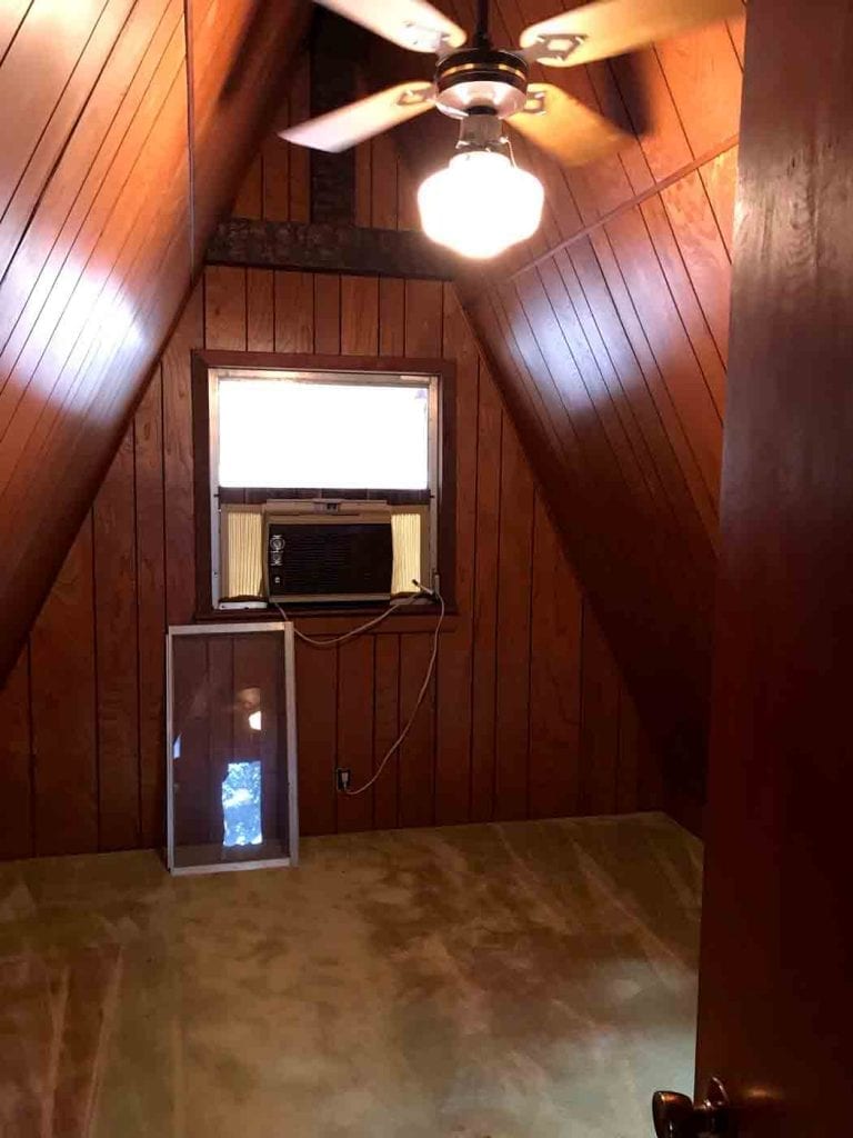 Interior of a rustic attic room with sloped wood paneling, showing a rectangular dormer window fitted with an air conditioner, ideal for a custom stained glass insert.