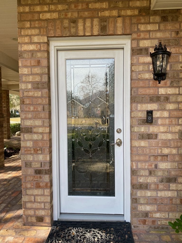 A white front door featuring a rectangular clear glass panel with an intricate floral and scroll design made from textured and beveled glass, set against a brick wall.