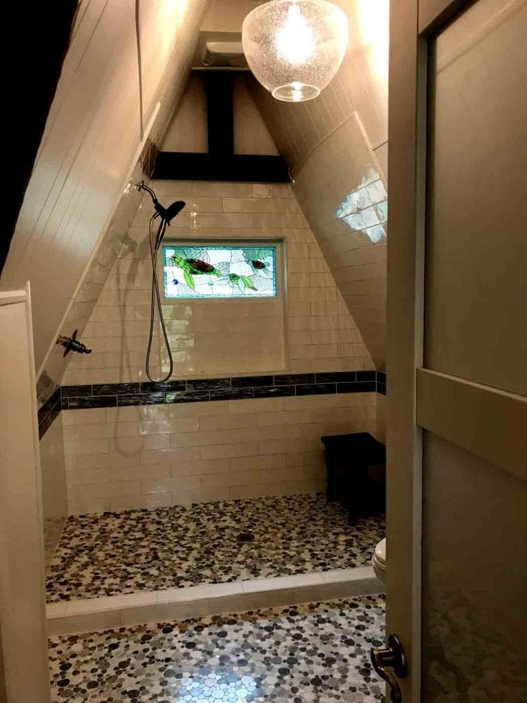 A rectangular stained glass window featuring two vibrant green sea turtles swimming in clear and light blue textured glass, installed centrally in a tiled A-frame shower. The shower has white subway tiles, a dark accent border, a pebble mosaic floor, and a black shower head. The unique A-frame ceiling is also tiled.
