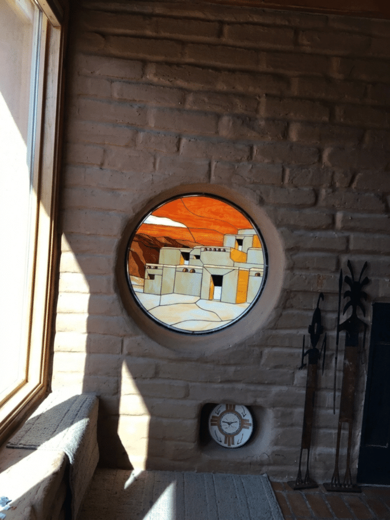 A circular stained glass panel depicting an adobe-style village with an orange sky and mountains, set into a mud brick wall.