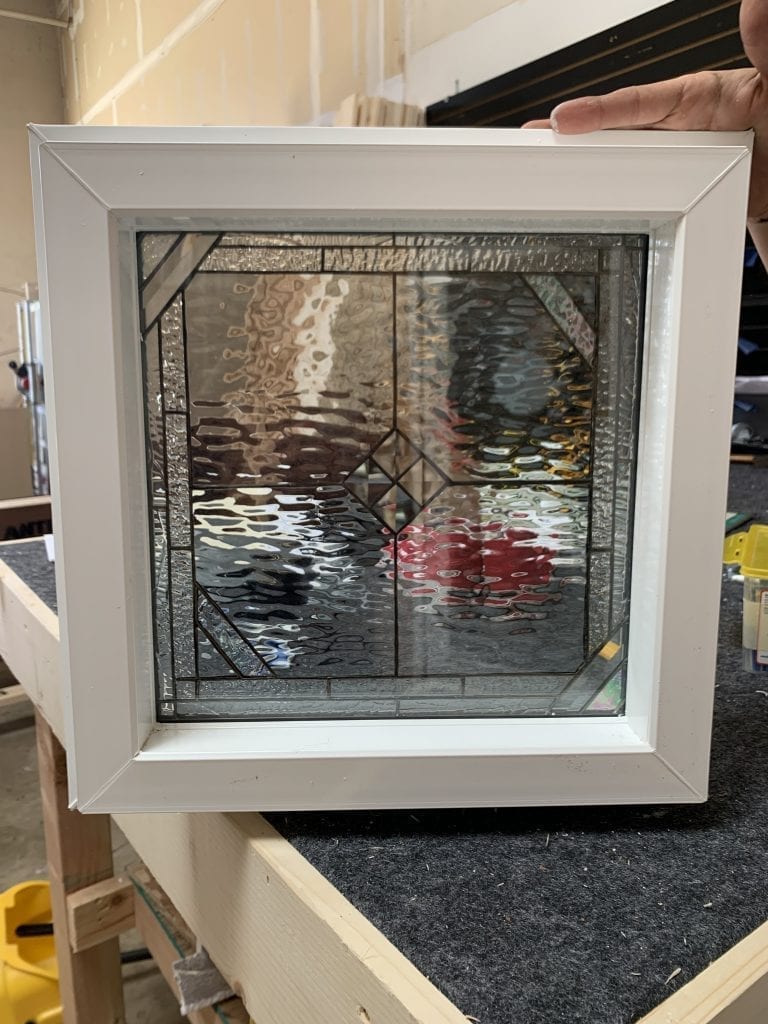 A square, white vinyl-framed window unit containing a leaded stained glass panel with a clear geometric pattern of textured and beveled glass, featuring a prominent central diamond motif.