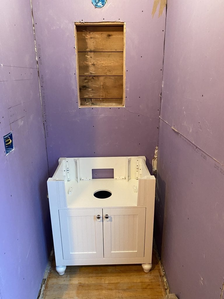 A bathroom under construction, featuring purple mold-resistant drywall, a white freestanding vanity cabinet base, and an empty recessed wooden wall niche, awaiting custom finishes.