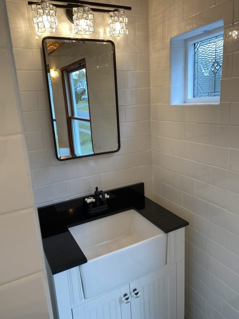 A rectangular leaded stained glass window, featuring a central clear glass bird design (resembling a dove or swallow) within a diamond motif, surrounded by various clear textured and beveled glass pieces, installed in a white frame on a white subway-tiled bathroom wall.