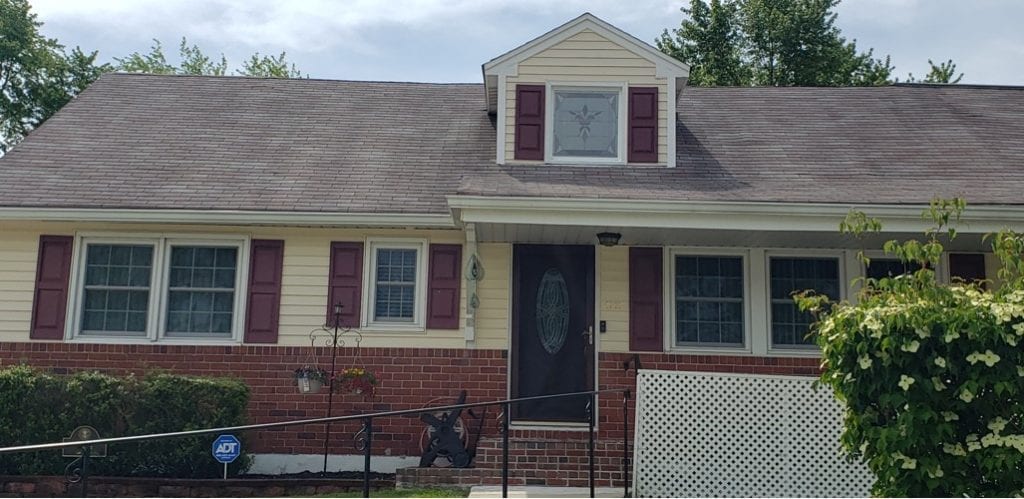 Exterior view of a yellow house with a brown roof, featuring a prominent dormer window with a clear geometric beveled stained glass panel, and a dark front door with an oval clear leaded glass insert. Maroon shutters flank the dormer and other windows, with a red brick base below.