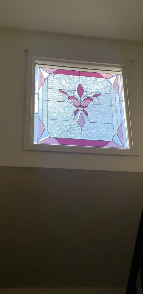 A rectangular stained glass window panel with a central stylized fuchsia and light pink floral or fleur-de-lis design. The motif is surrounded by clear textured glass and framed by fuchsia and light pink geometric border elements, all set within a white window casing.