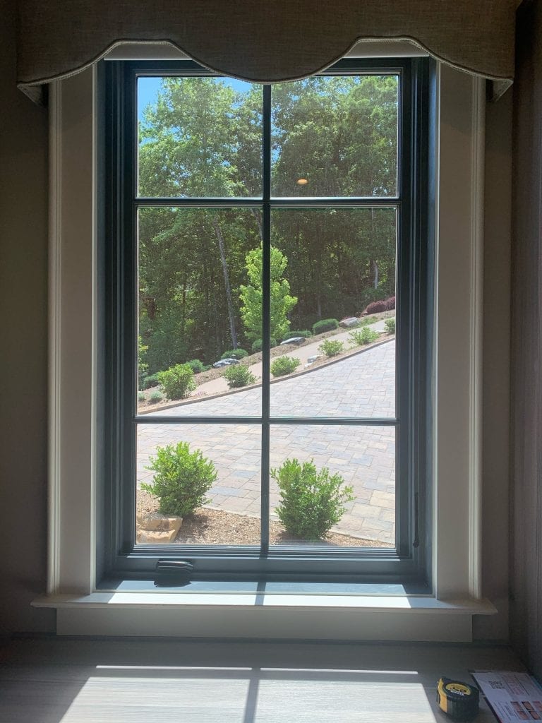 A tall rectangular window with dark muntins creating a six-pane grid, looking out onto a sunny, green outdoor scene with a paved driveway, bushes, and mature trees. A tan fabric valance tops the window, and sunbeams highlight the white sill and a yellow tape measure on the desk.