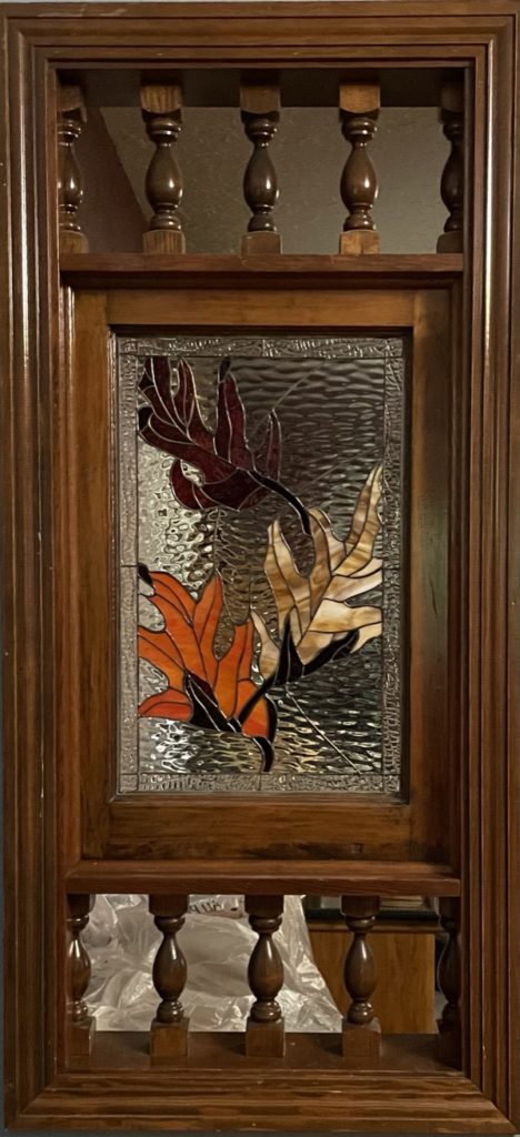A rectangular stained glass panel set within a dark wooden frame, featuring three autumn leaves in deep red, vibrant orange, and warm tan art glass against a clear, textured, leaded glass background. The wooden frame is accented with decorative turned spindles above and below the stained glass section.