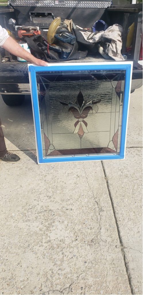 A rectangular leaded stained glass panel in a white frame featuring a central dark and light brown fleur-de-lis design against a clear textured glass background.