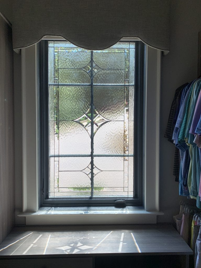 A rectangular leaded glass window panel with clear textured and beveled glass, featuring two central diamond designs and smaller geometric bevels. The panel is set in a dark frame within a white window trim, topped by a light grey valance, and sunlight casts patterns on a surface below.