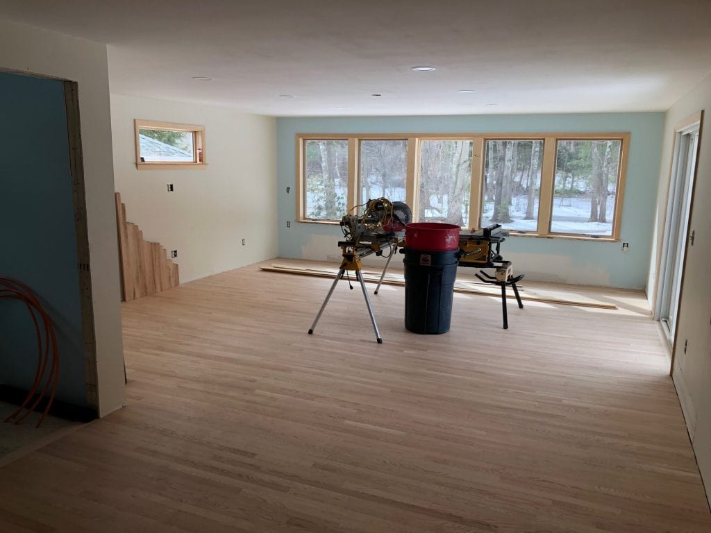 An unfinished but newly renovated living room featuring pale hardwood floors, light cream walls, and a light blue accent wall around large windows. Construction equipment, including a miter saw and a red and black shop vac, is in the center. Through the windows, a snowy winter landscape with bare trees is visible.