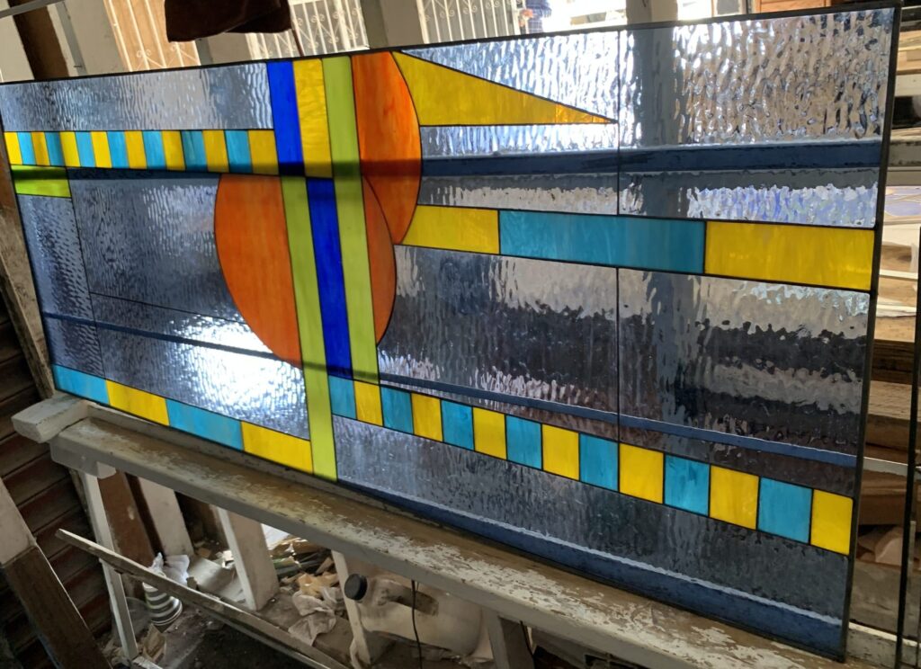 Rectangular leaded stained glass panel featuring an abstract geometric design with a prominent orange disc, intersected by vertical blue and green lines, and surrounded by horizontal bands of yellow and teal rectangles. The background is a textured, dark clear glass, all set in black leading.