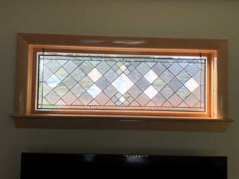 A rectangular leaded stained glass panel with a repeating geometric diamond pattern, featuring clear textured and beveled glass, accented with subtle hints of light blue and amber. The panel is set in a light wood frame, installed as a transom window above a television.