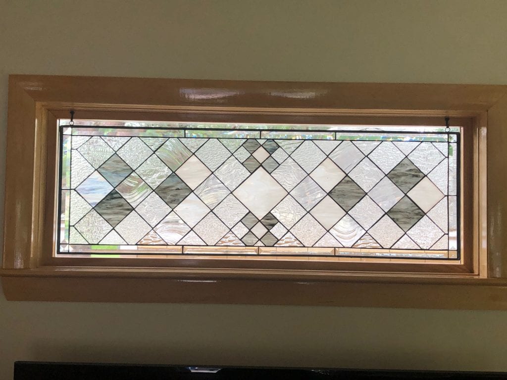 Rectangular clear and grey stained glass transom panel with a repeating geometric diamond pattern. The panel incorporates various textures of clear glass, opaque white glass, and streaky grey glass, all connected by black lead lines. It is suspended within a natural wood window frame, offering privacy and light.
