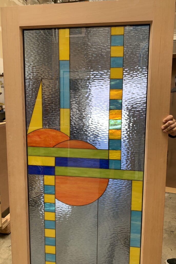 A large rectangular stained glass panel set within a natural wood frame, displaying a vibrant abstract geometric design. The panel features a background of clear, rippled textured glass, punctuated by vertical columns of alternating yellow and teal rectangles. Dominating the lower half are two large orange semi-circular shapes, intersected by prominent horizontal bands of green and blue glass. A sharp yellow triangular shape appears in the upper left, contributing to the panel's dynamic and contemporary aesthetic.