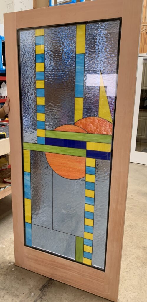 An abstract geometric stained glass door panel with a vibrant design in blue, yellow, orange, and green, featuring a large orange textured semicircle and clear textured glass, set in a natural wood frame.