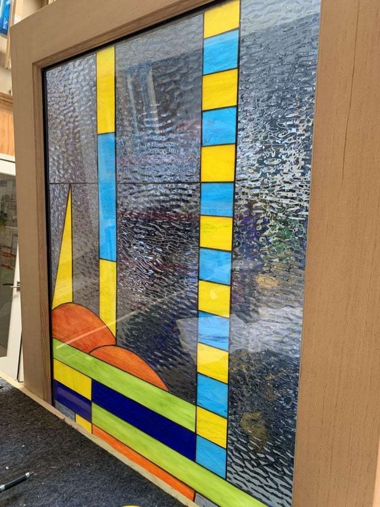 A rectangular stained glass window panel set in a light wood frame, showcasing a vibrant abstract geometric design. Large sections of clear, rippled textured glass are interspersed with vertical and horizontal segments of bright yellow, sky blue, lime green, and dark blue. Abstract, rounded orange shapes resembling hills appear at the bottom left of the panel.