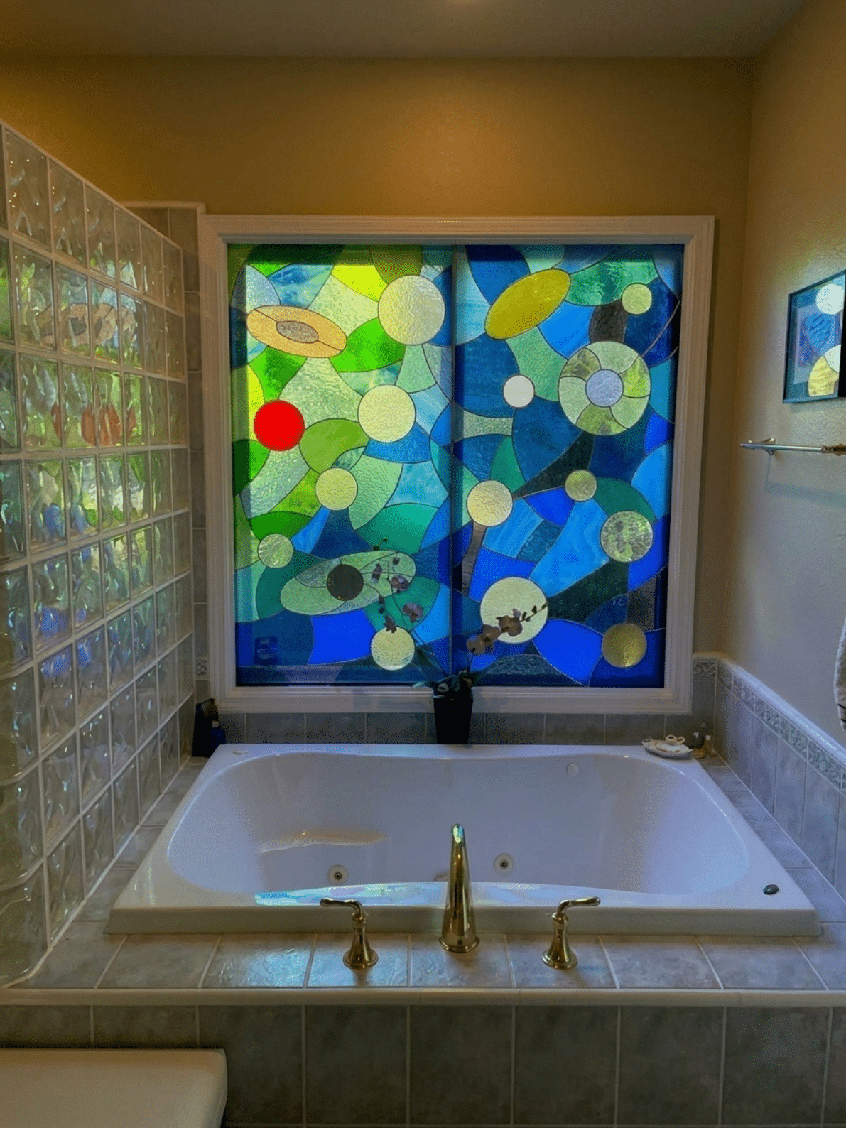 A rectangular, leaded stained glass window composed of two vertical panels, featuring an abstract design of various sized circles and organic shapes in shades of blue, green, and yellow, with accents of red and gold. The window is installed above a white bathtub in a bathroom.