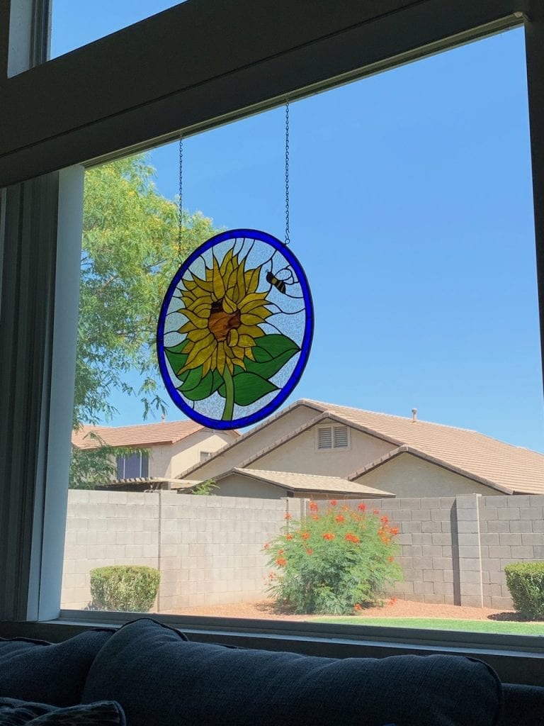 A circular stained glass suncatcher depicting a vibrant yellow sunflower with a brown center, green leaves, and a small black and yellow bee flying near its petals. The suncatcher has a royal blue border and hangs by two metal chains in front of a clear window, showcasing a bright blue sky and distant residential buildings with a beige stucco wall and green bushes in the background.