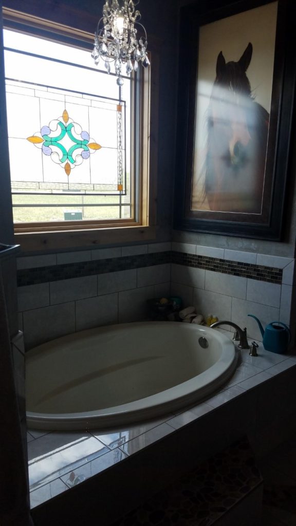 A rectangular stained glass window panel installed in a bathroom, positioned above a white bathtub. The panel features a central symmetric geometric design with teal, light purple, and amber glass accents, framed by clear textured leaded glass. A small crystal chandelier hangs in the foreground, and a large framed horse portrait is visible on the adjacent wall.