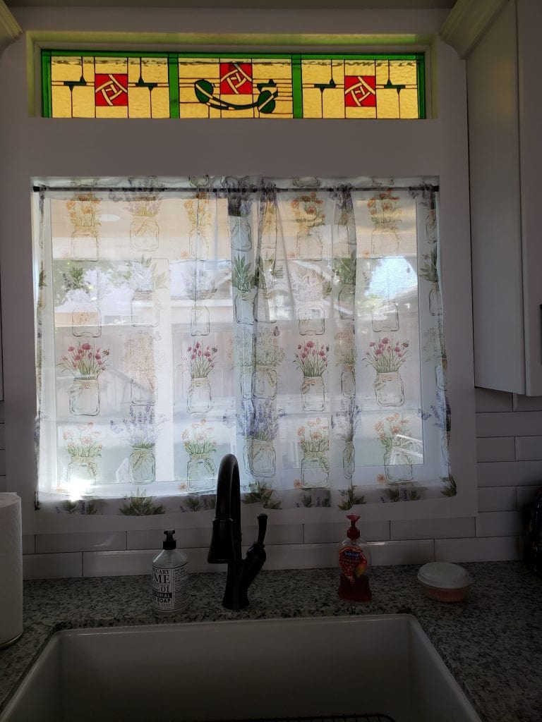 A rectangular stained glass transom window featuring a geometric Art Nouveau-inspired design. The panel has a prominent yellow-gold glass background, framed by vibrant green leaded lines, and interspersed with striking red squares containing intricate, interwoven abstract patterns. It is installed above a kitchen window with patterned curtains, a sink, and white subway tile backsplash.