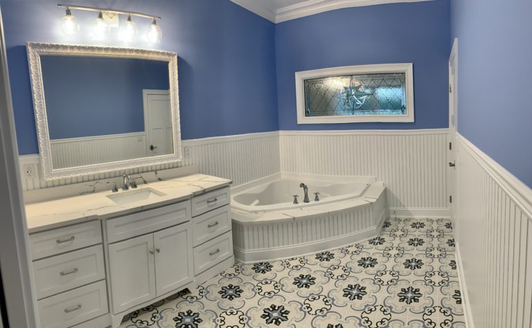 A horizontal clear beveled stained glass transom window featuring a central multi-faceted compass rose or starburst design, surrounded by various textured clear glass panels. The window is installed above a corner bathtub in a blue and white bathroom.