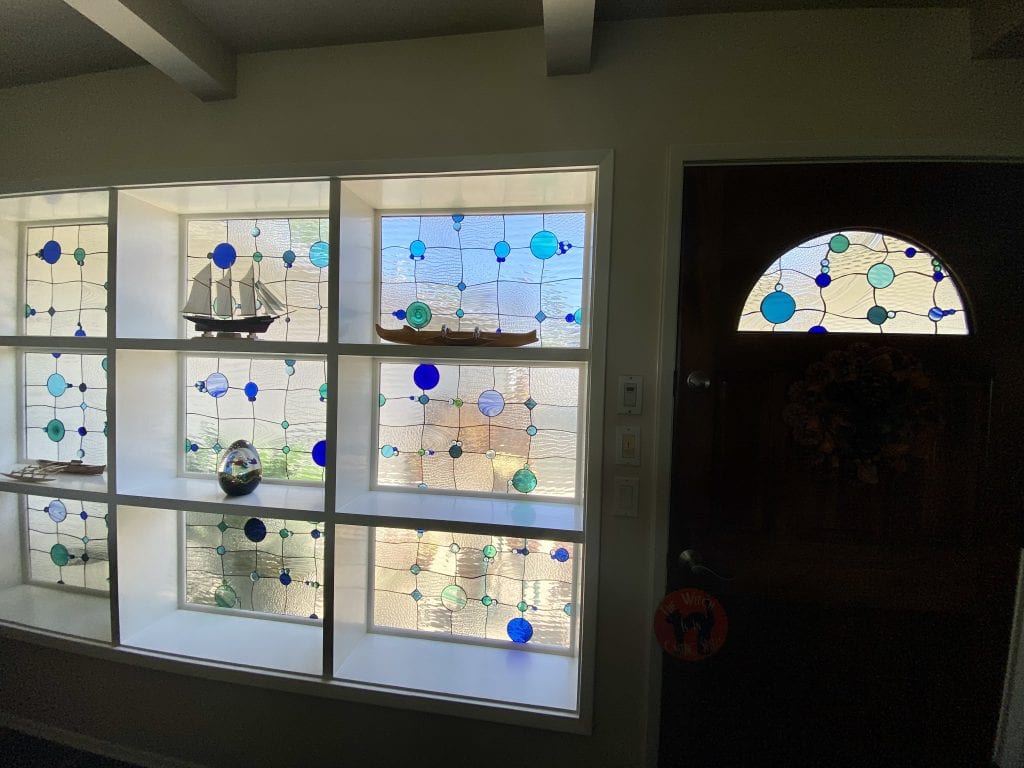 A contemporary custom art glass installation featuring a large, multi-panel window unit and a matching arched door transom. Each panel consists of clear textured glass with an abstract pattern of various sized blue and teal circular and square glass jewels connected by dark lead lines. Decorative items like a model sailboat and kayak are placed on the window sills. The overall design evokes an underwater or bubble motif, enhancing light and privacy.